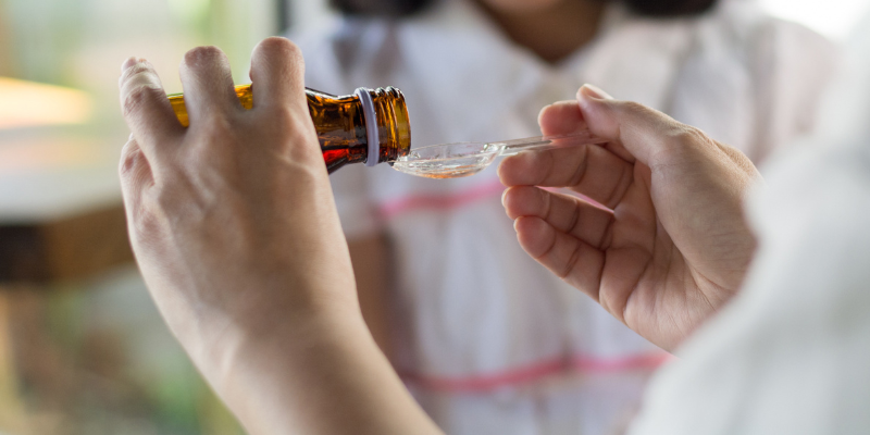 Child girl smiling want to take medicine, hands of mom pouring cough syrup medicine into clear spoon to daughter